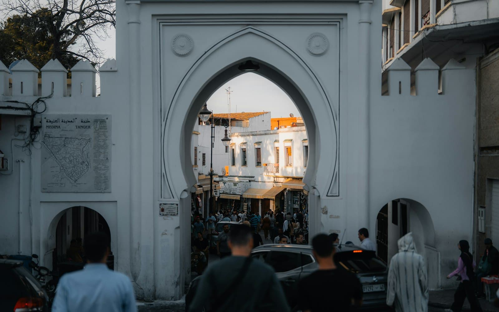 Bab Fahs, la grande porte blanche crénelée entre le Grand Socco et la médina de Tanger, avec la plaque « Medina Tanger » et des passants de dos