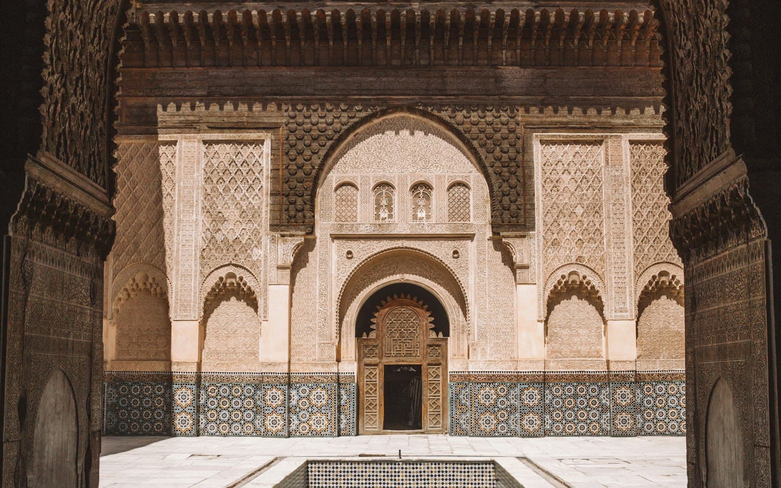 Cour de la Medersa Ben Youssef à Marrakech : façade en stuc sculpté, porte en bois, zelliges et bassin