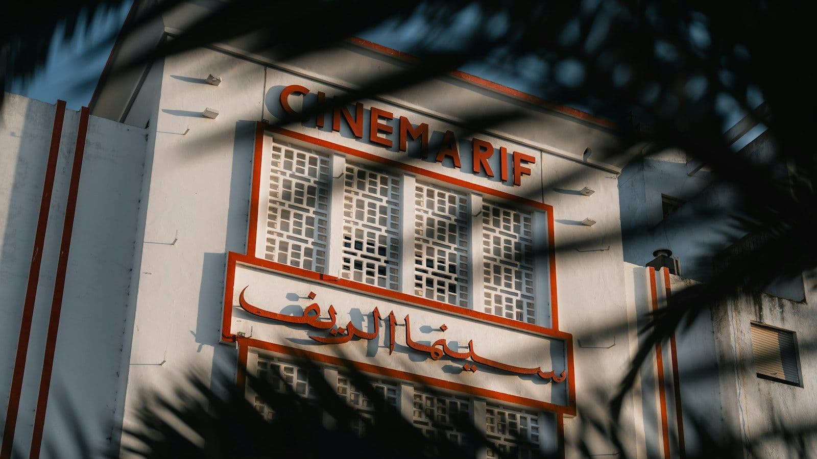 La façade Art déco blanche et rouge du Cinéma Rif à Tanger, siège de la Cinémathèque, vue à travers des feuilles de palmier
