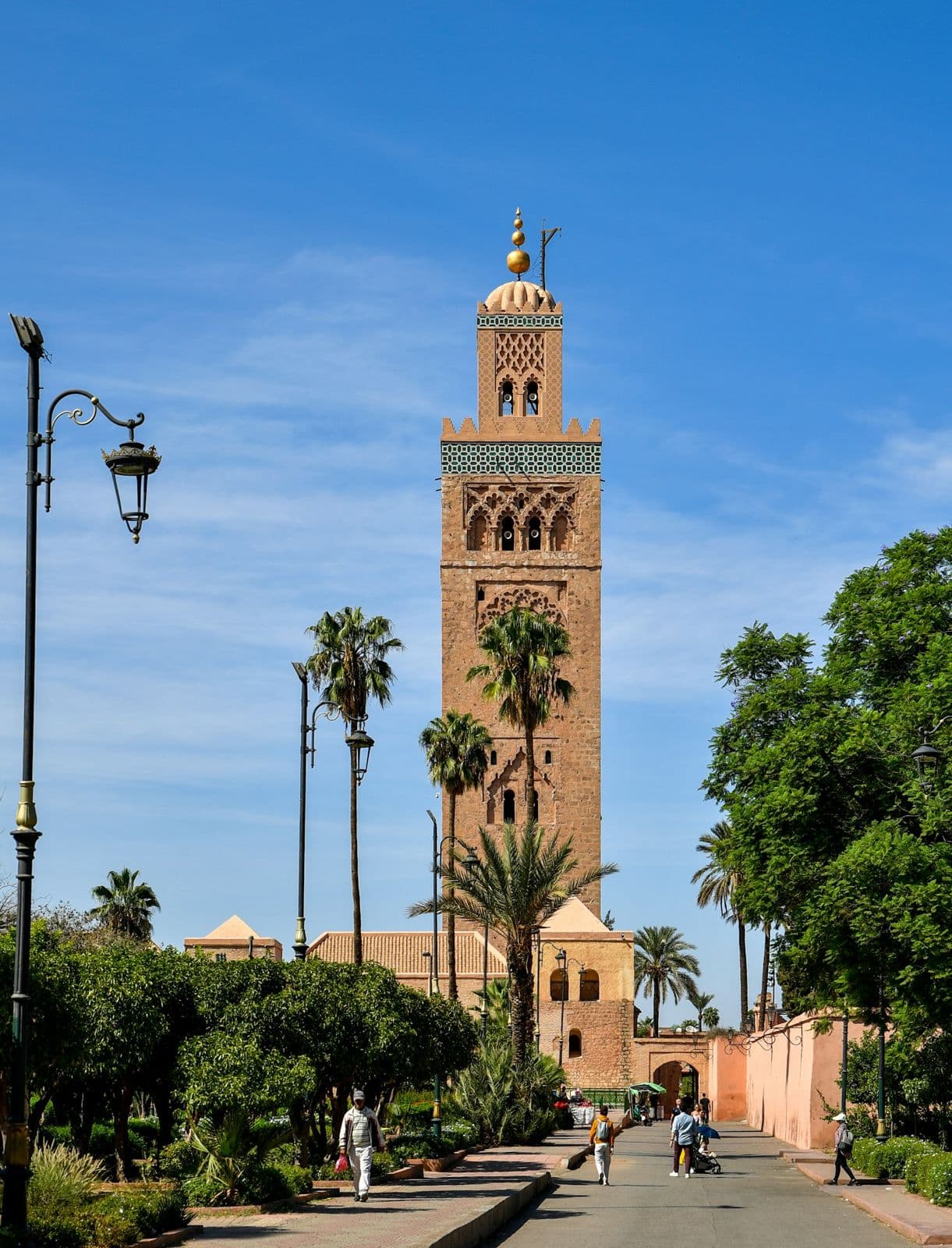Le minaret de la Koutoubia à Marrakech vu depuis une allée bordée de palmiers, sous un ciel bleu, avec quelques passants