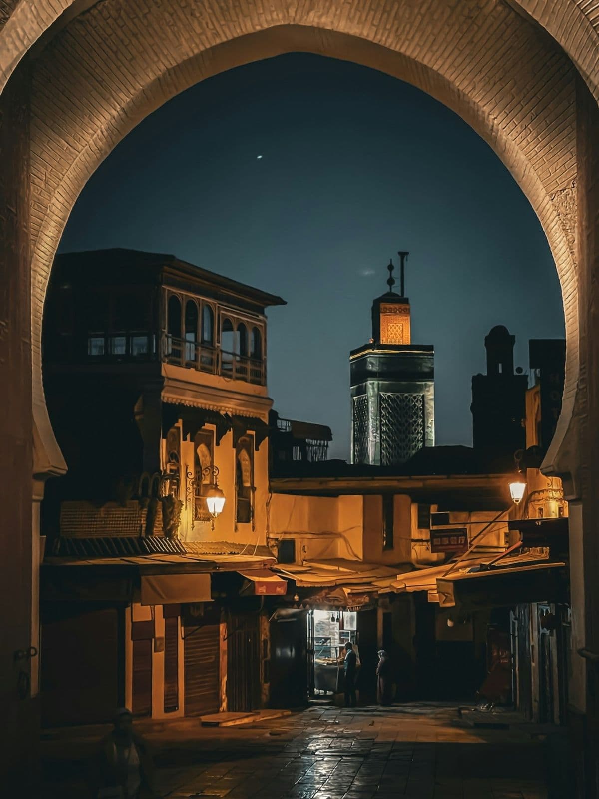 Vue de nuit à travers une grande porte en arc de médina marocaine : un minaret éclairé, des façades à balcons de bois et des rideaux de boutiques baissés