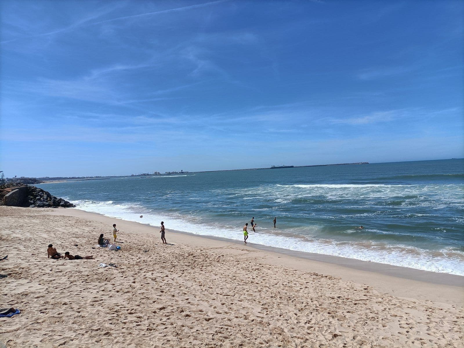 Plage de sable à Mohammédia sous un ciel bleu, avec quelques promeneurs au bord de l'eau et une jetée à l'horizon