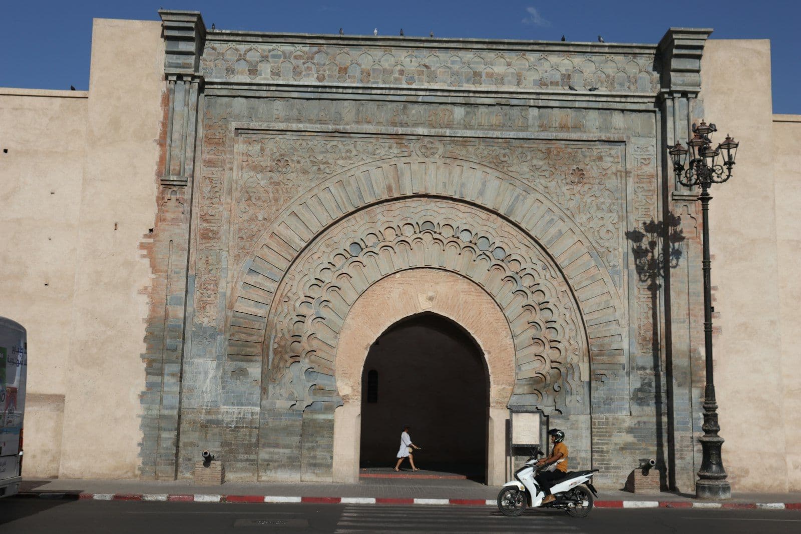 Porte monumentale sculptée dans les remparts ocre de Marrakech, sous un ciel bleu, avec un scooter qui passe devant