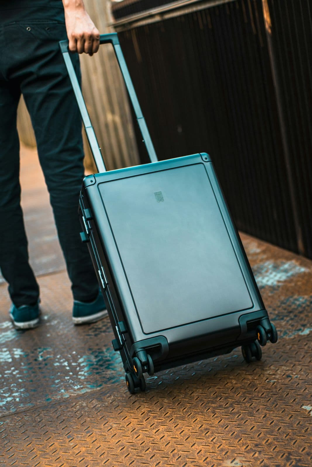 Voyageur tirant une valise rigide grise à roulettes sur un sol en tôle striée, vu de dos à hauteur des jambes