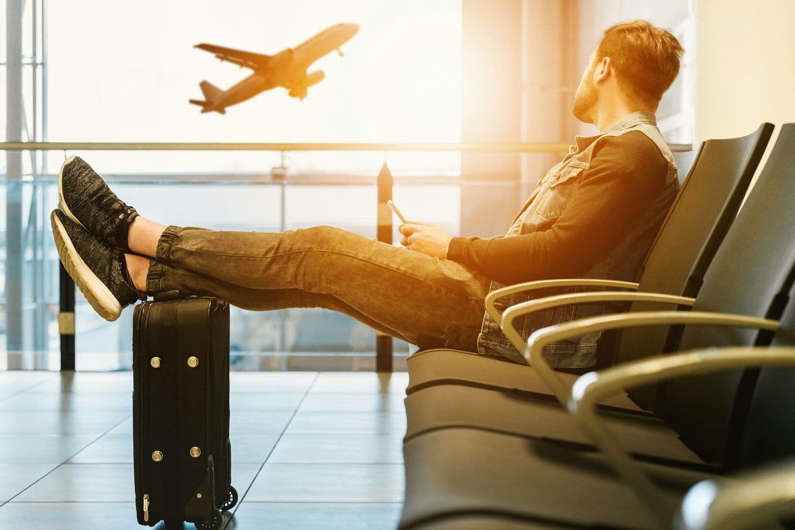 Voyageur assis dans une salle d'embarquement, les pieds posés sur sa valise, regardant un avion décoller derrière la baie vitrée