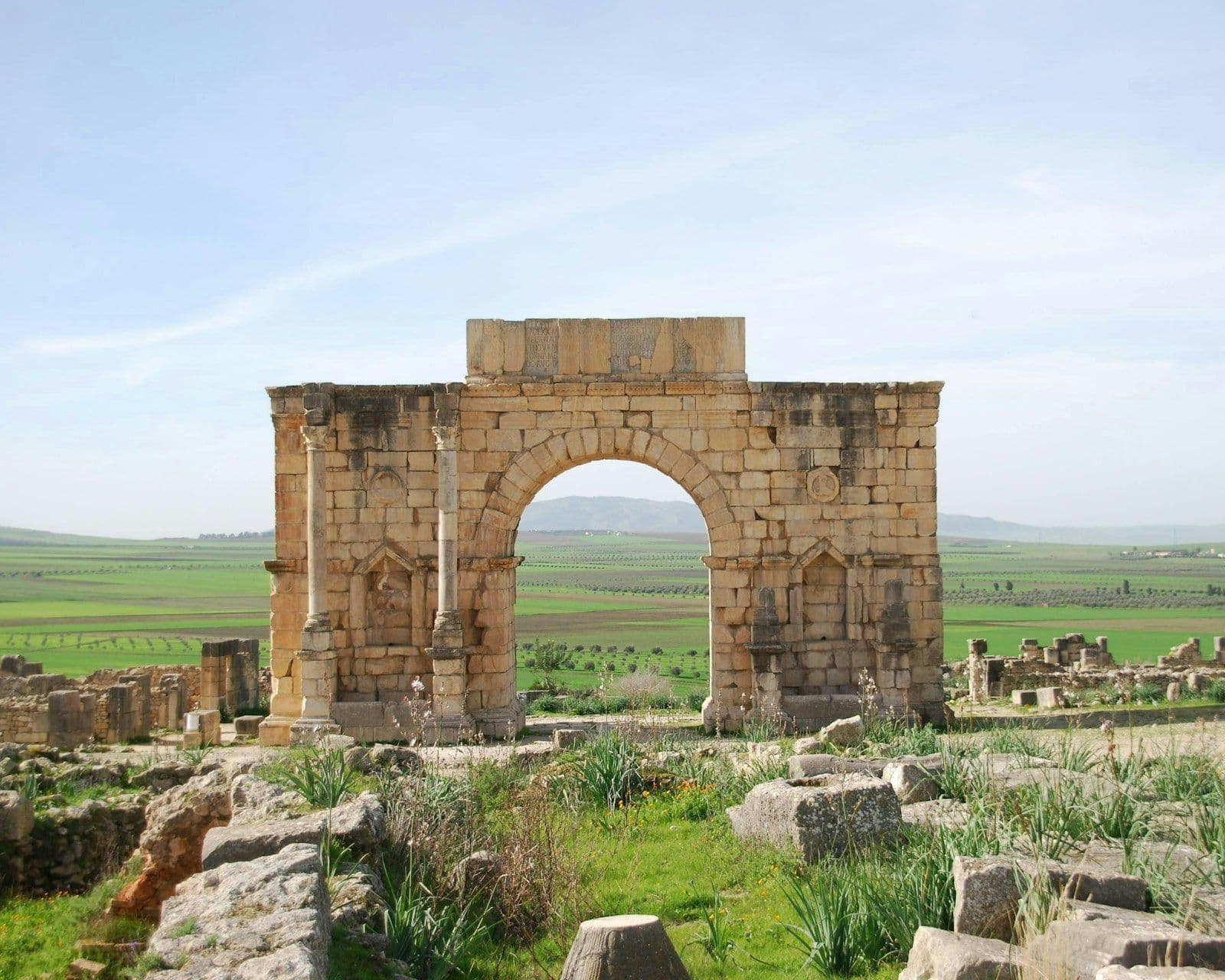 Arc de triomphe romain en pierre ocre au milieu des ruines de Volubilis, avec des champs verts et des collines en arrière-plan