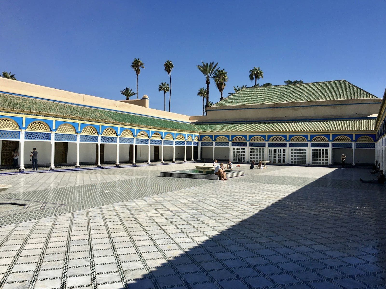 La grande cour pavée de marbre du palais de la Bahia à Marrakech, entourée de galeries aux boiseries bleues et de toits de tuiles vertes, sous un ciel bleu