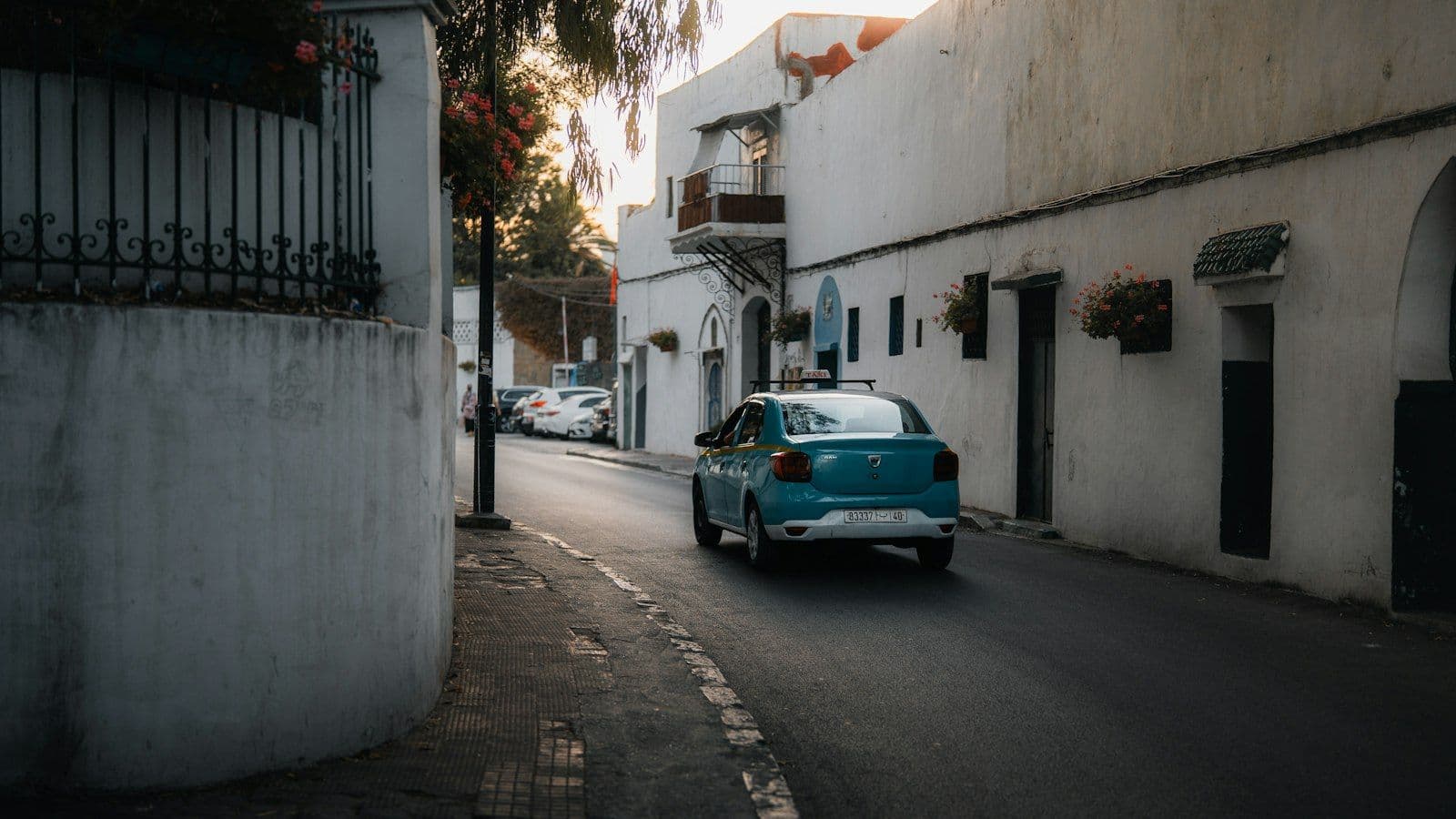 Un petit taxi bleu turquoise roule dans une rue bordée de maisons blanches, en fin de journée