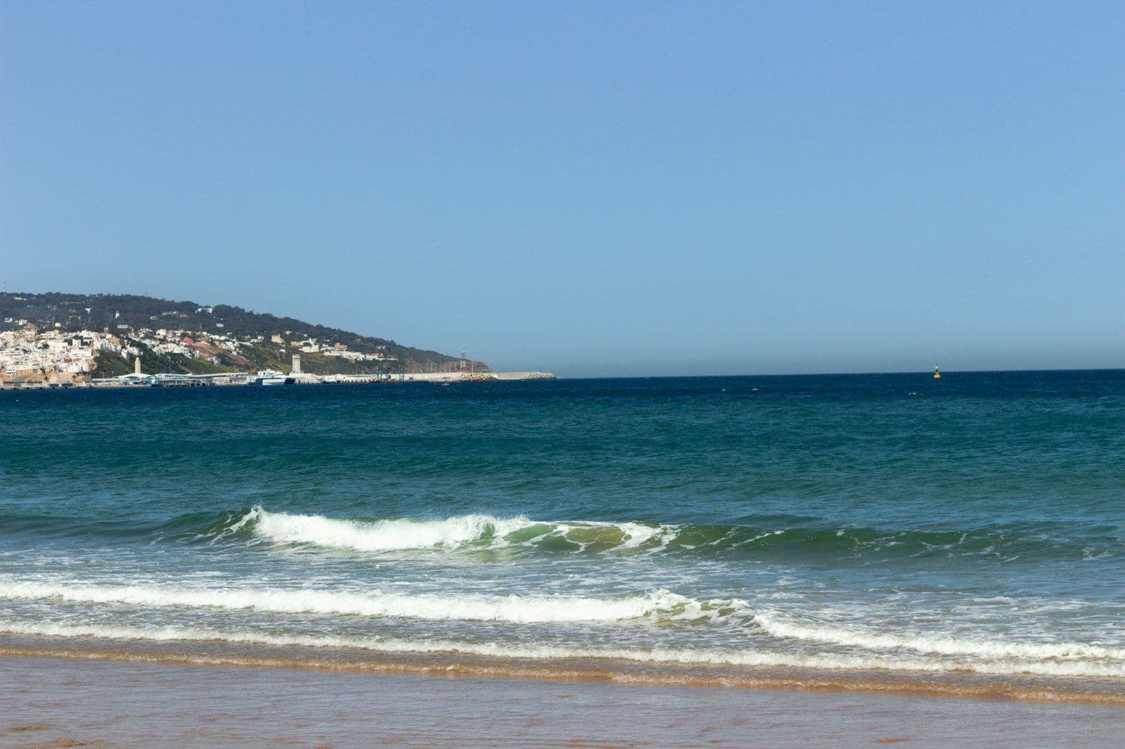 Vagues sur une plage de la baie de Tanger, avec la ville blanche et le port sur la colline en arrière-plan, sous un ciel bleu
