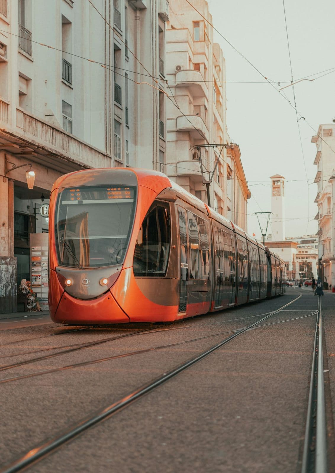 Un tramway orange circule sur ses rails dans une rue du centre de Casablanca, entre des immeubles anciens, avec une tour d'horloge au fond.