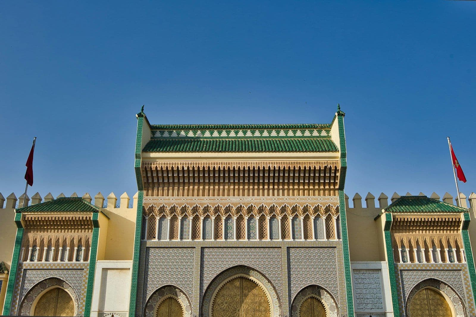 Les portes dorées du Palais royal de Fès (Dar el-Makhzen), sous un ciel bleu, avec leurs toits de tuiles vertes et deux drapeaux marocains