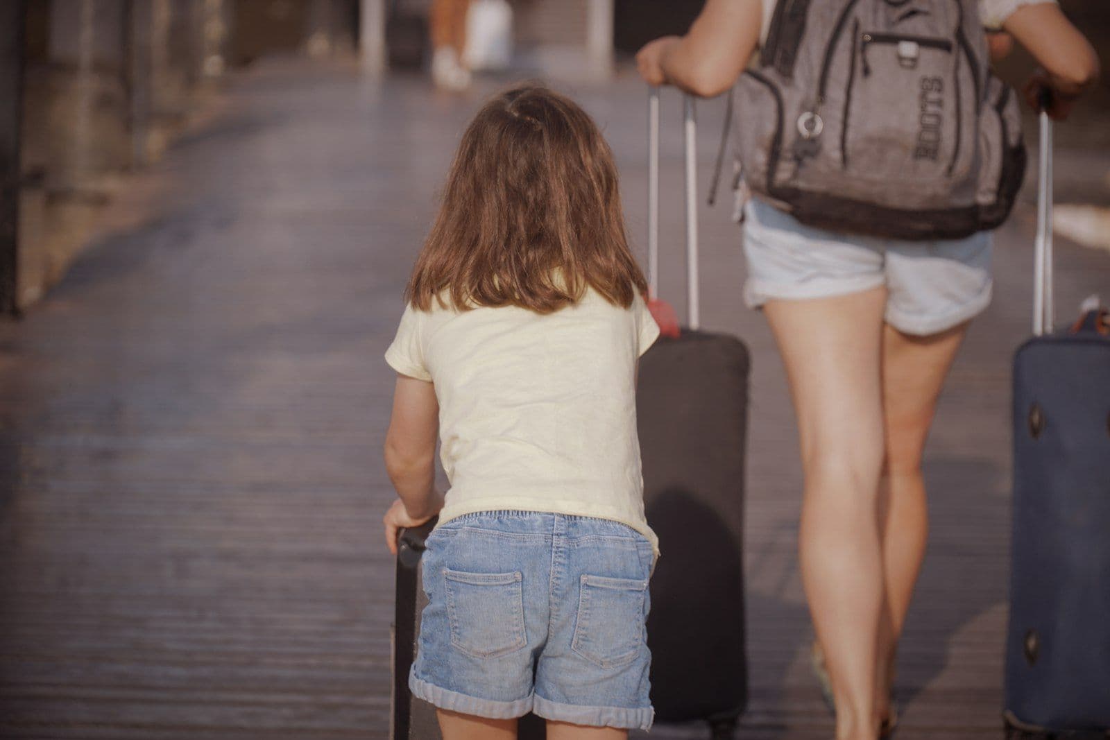 Une enfant et un adulte, vus de dos, tirent leurs valises à roulettes sur un passage en bois